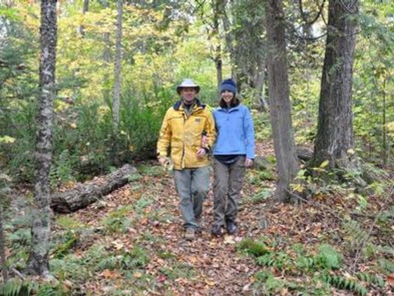 A man and woman that are arm in arm in the woods. There are orange and brown leaves on the ground along with ferns. There are trees behind the couple. 
