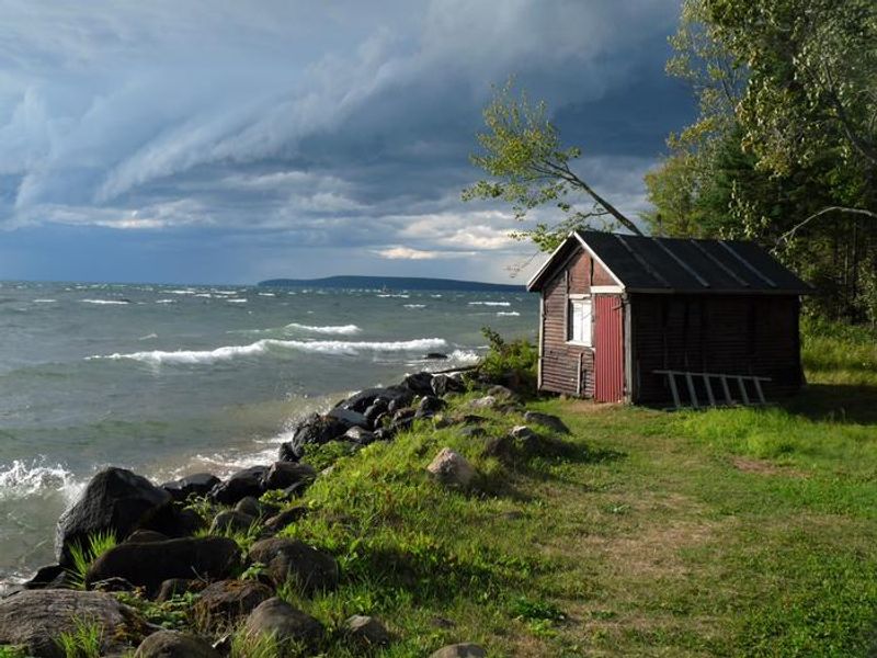A color photo with a dark blue ominous sky with clouds and choppy lake conditions. On the left side of the photo are white cap waves heading toward the rocky shoreline and land. There is a small dark red/brown older building on the right side sitting on the grounds of Manitou Island. 