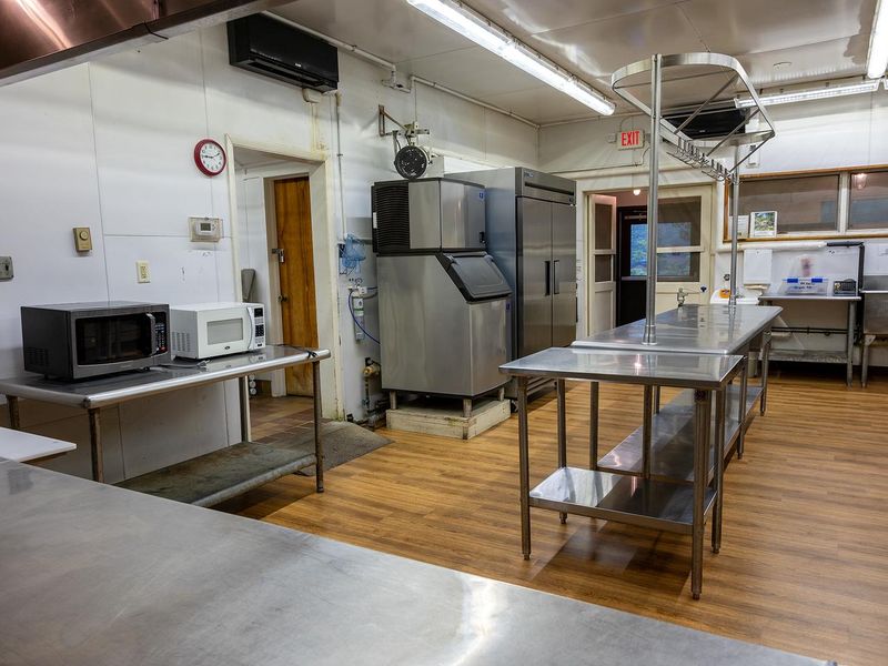 Dining hall front door view of the kitchen. Stainless serving line with 3-well hot food table, prep tables, ice machine, prep refrigerator, microwave and double sink.  Not visible are 2 large storage areas, 1 small bathroom, walk-in refrigerator, and double freezer.
