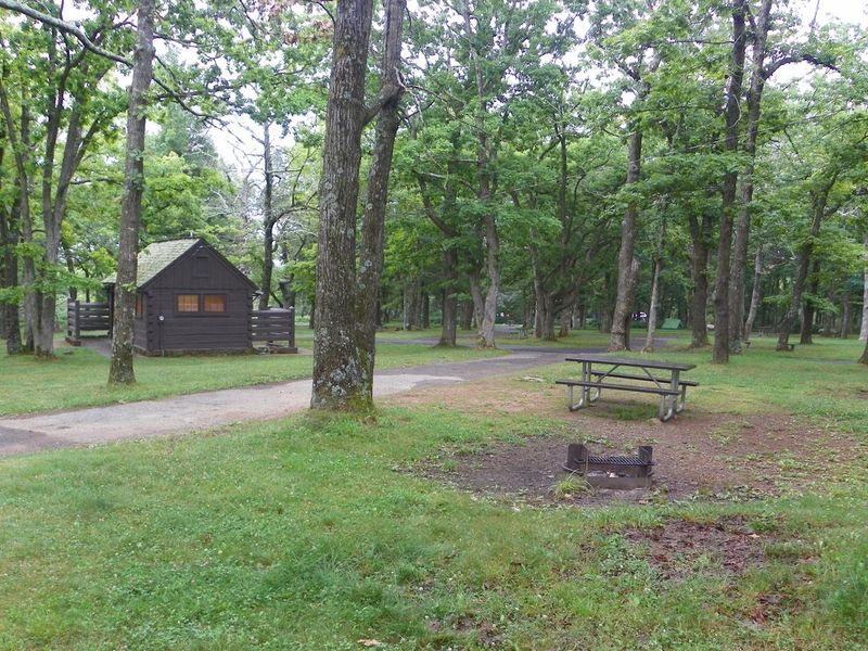 The driveway of a pull through campsite passes between tree trunks. At right, a picnic table and fire ring surrounded by a green lawn. In the background, is a wood cabin restroom building 