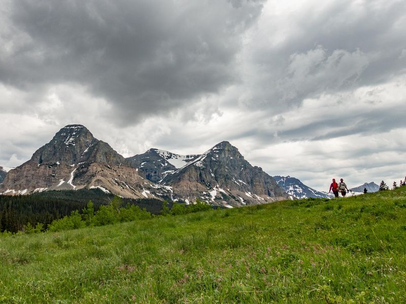 Two people walk down a trail near the Cutbank Campground under cloudy skies. Rocky snow capped peaks rise in the background and the foreground is filled with thick grass.