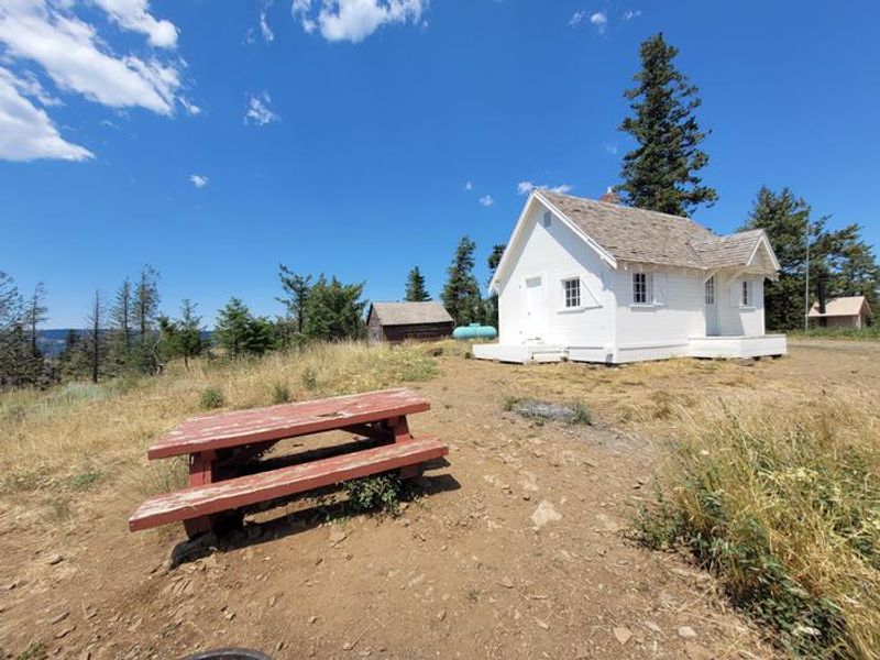 Outside the Wenatchee Guard Station is equipped with two porches, a picnic table, firepit, and vault toilet. Picture taken before updates in summer of 2023.