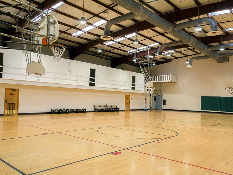 Interior of gym.  Wooden marked basketball court with 4 backboards/nets and 4 benches on sidelines. Metal beam ceiling with skylights. 3 doors visible to upper classrooms. Women's and Men's bathrooms are accessible from the gym court.