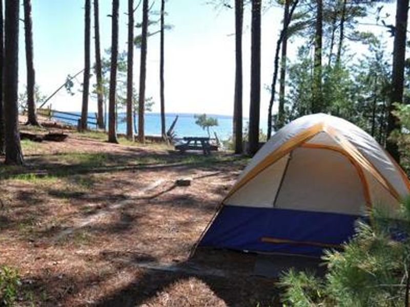 A campsite with a lake view in the background. There is a small individual tent on the right side of the photo. A picnic table is centered in the middle of the photo. There are trees surrounding the tent and scattered on the left side of the photograph. 