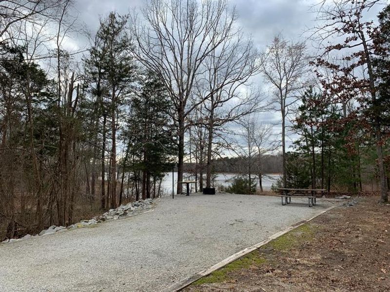 This is a photo of site 6. Most campsites are graveled. This site has a picnic table, utility table, lantern post and a fire ring/grill. 