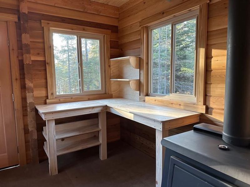 Interior of cabin kitchen area consisting of two wall shelves, a corner countertop and two shelves below the countertop. Wood stove to the right of the counter. 