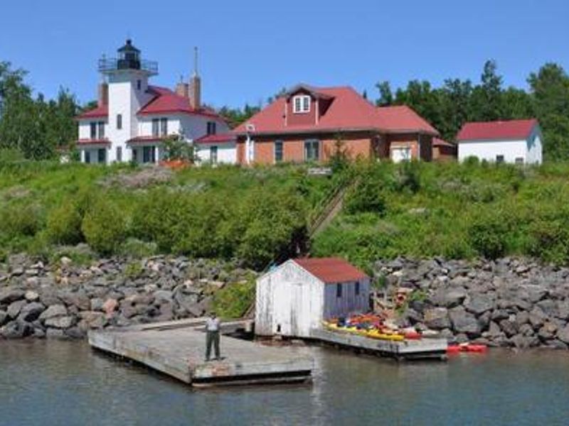 A ranger standing on the left side of the U-shaped dock with a boat house in the off-centered of the U. A lighthouse and other building on a bluff behind the dock. 