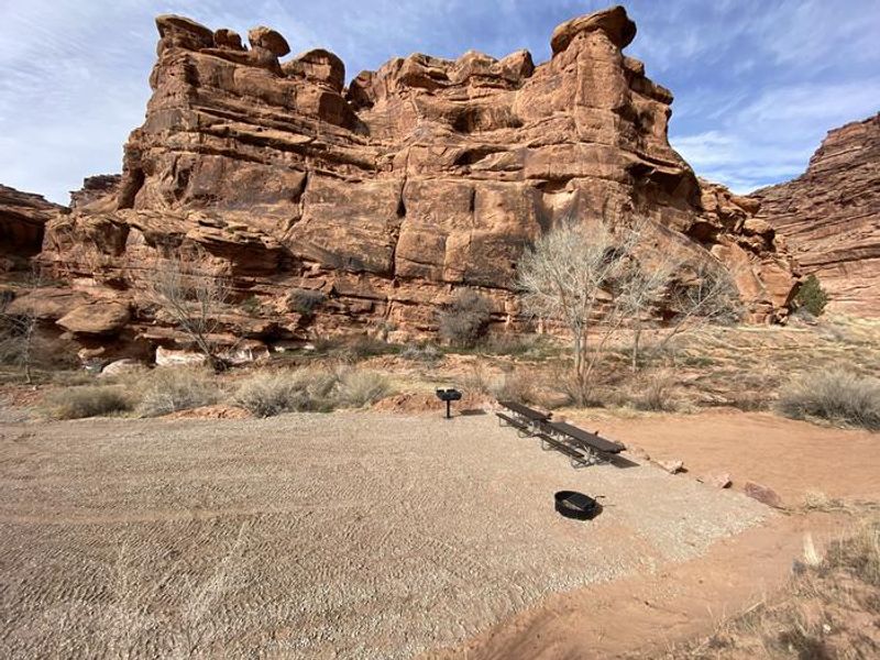 Group campsite with picnic tables, fire pit, and grill, cliffs in background.