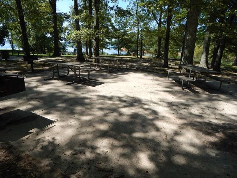 In this picture there are accessible picnic tables and grills. They are on a concreted pad with an accessible restroom near by. These are located near the OSAGE picnic shelter. 