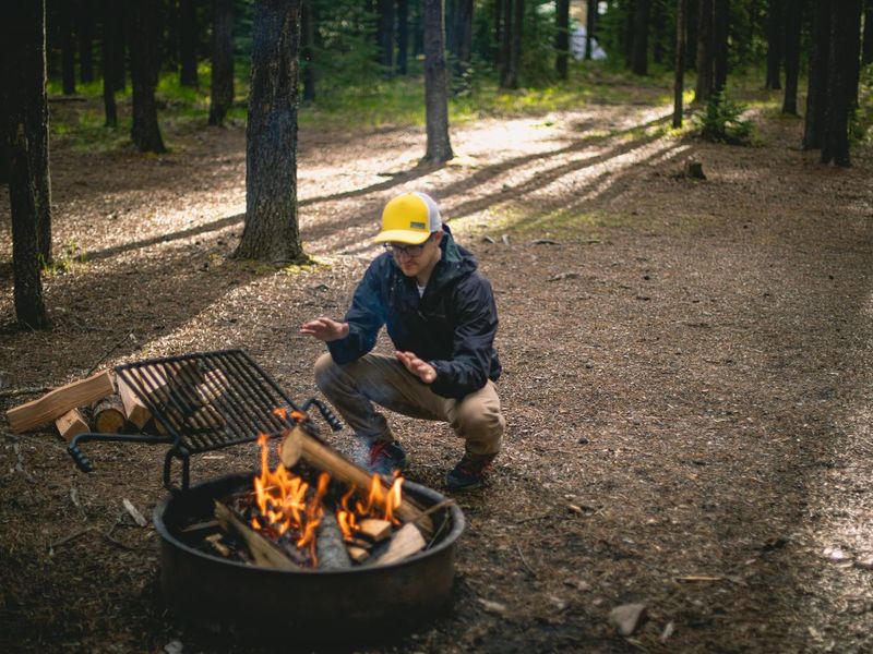 A camper wearing a bright hat warms their hands while squatting near a campfire in a forest clearing.
