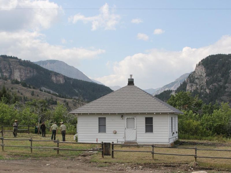 Small, white cabin surrounded by mountains, trees, and people