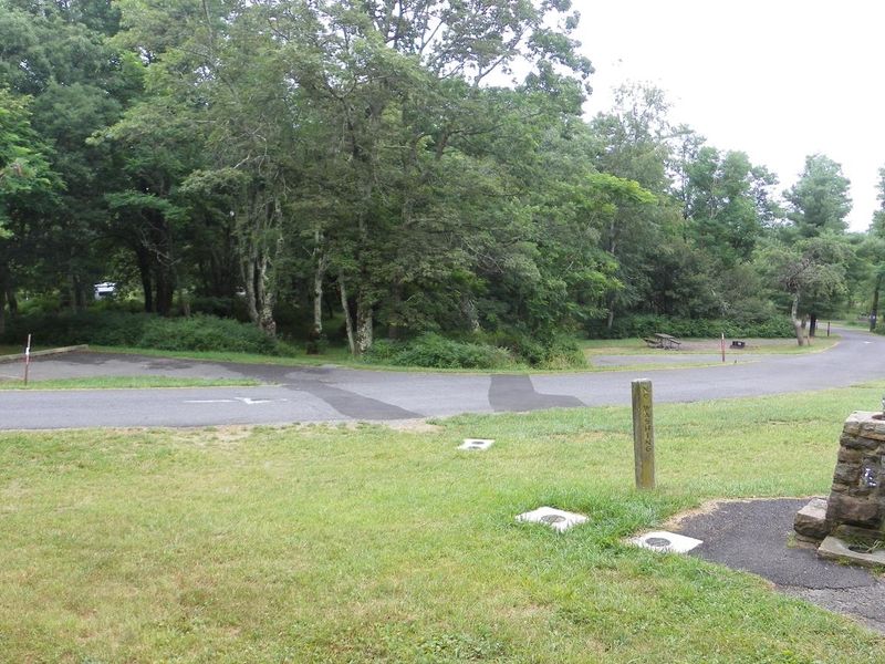 A stone water fountain next to a road that is lined with campsites. Each campsite is surrounded by trees and bushes.