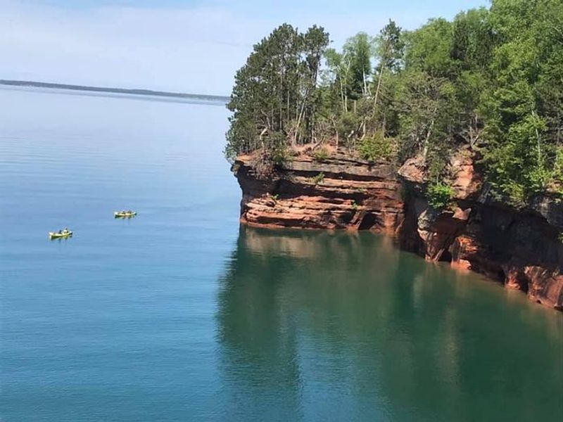 Kayakers sits in sea kayaks on calm waters. There is a blue sky to the left and brownstone cliffs to the right of the kayakers with some trees on top.