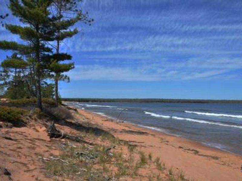 A landscape photo of a sandy beach with beach grass and trees on the left. The lake on the right side of the beach has white cap waves. 