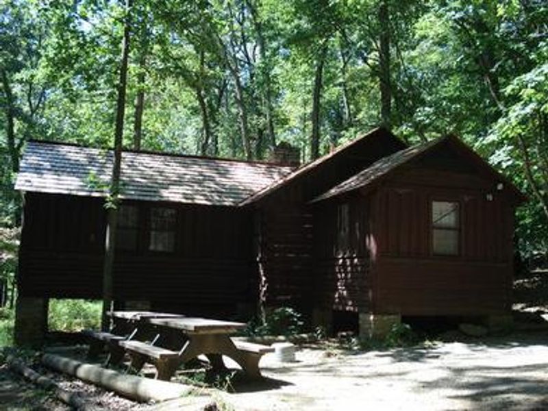American chestnut log cabin #16 with two picnic tables and outdoor grill/firepit surrounded by trees. Cabin #16 has electricity, a full bathroom, small refrigerator, microwave and heat.  The cabin can accommodate 6 individuals.
