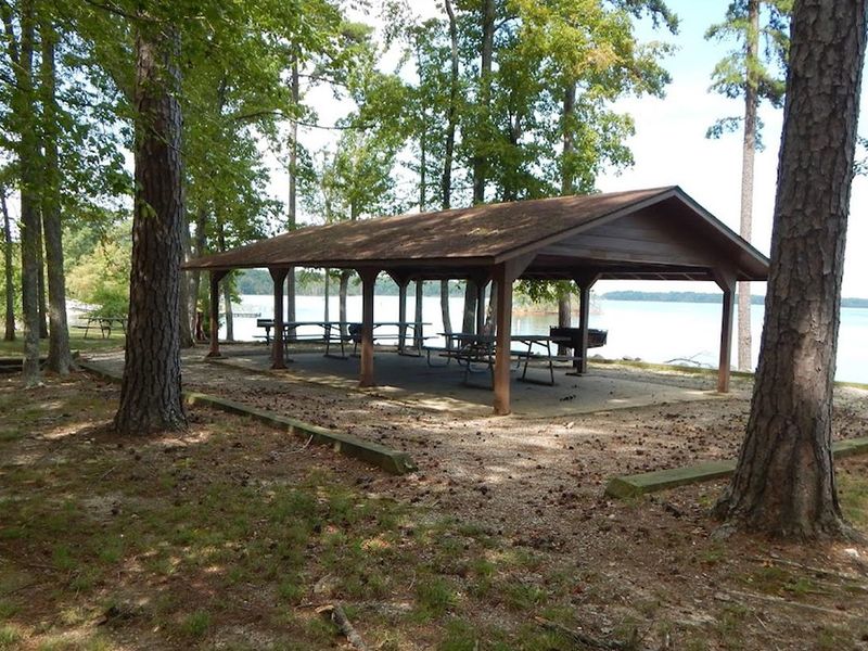 This is a picture of the Ivy Hill Shelter that is reservable. There are picnic tables under a covered shelter. This area is shaded but has a wonderful view of the lake. There are trash cans located throughout the park. 