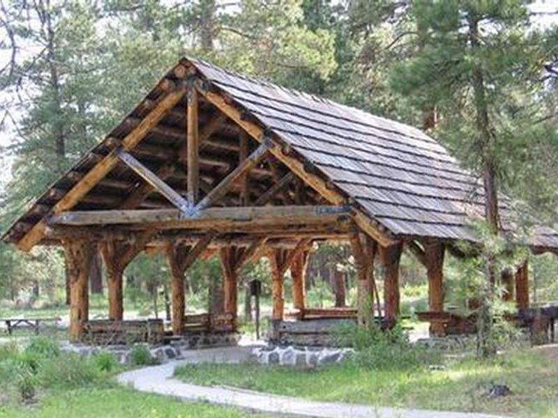 Large log framed picnic shelter next to pine trees and a grassy meadow accessed by a flat path. 