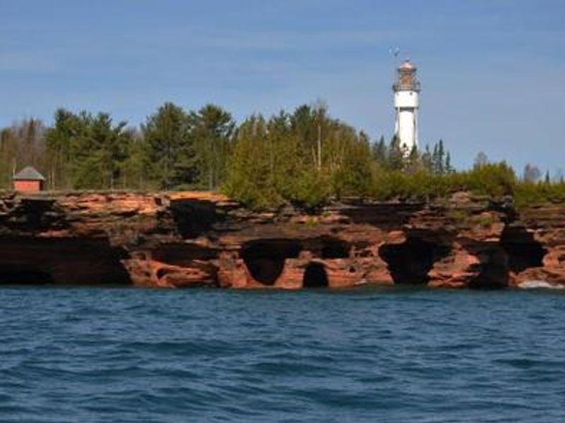 A landscape photo of water in the foreground with sea caves and a lighthouse in the background. The lighthouse sits behind green trees that are half the height of the lighthouse which are on the top of the sea caves. There is a small building on the top left side of the sea caves. 