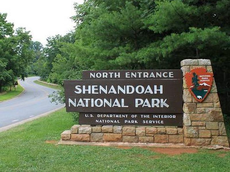 A narrow road winds between trees, past a stone post holding a wooden sign that reads "North Entrance Shenandoah National Park" with the National Park service arrowhead logo.