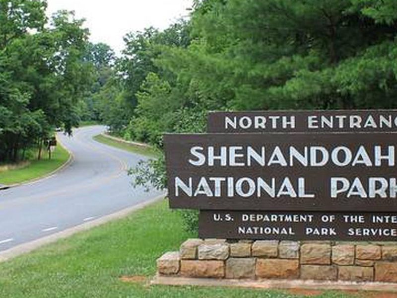 A narrow road winds between trees, past a stone post holding a wooden sign that reads "North Entrance Shenandoah National Park" with the National Park service arrowhead logo.