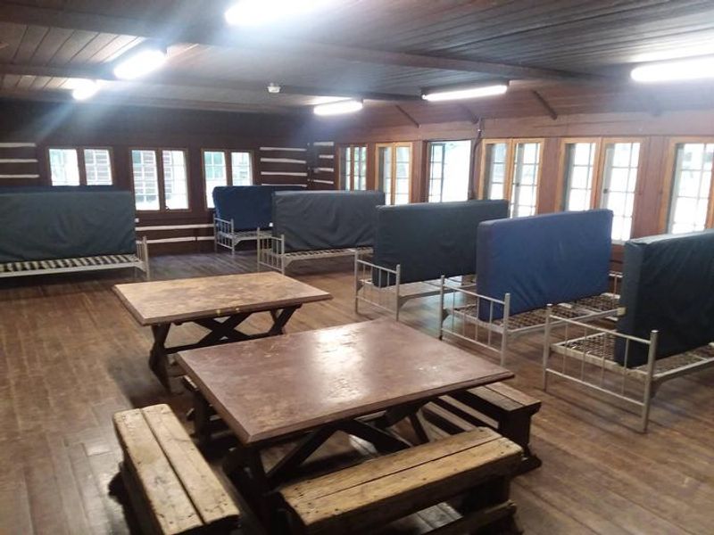 Interior of a Camp Misty Mount lodge. 2 square tables and 4 benches.  8 single metal cots with vinyl covered mattresses.  