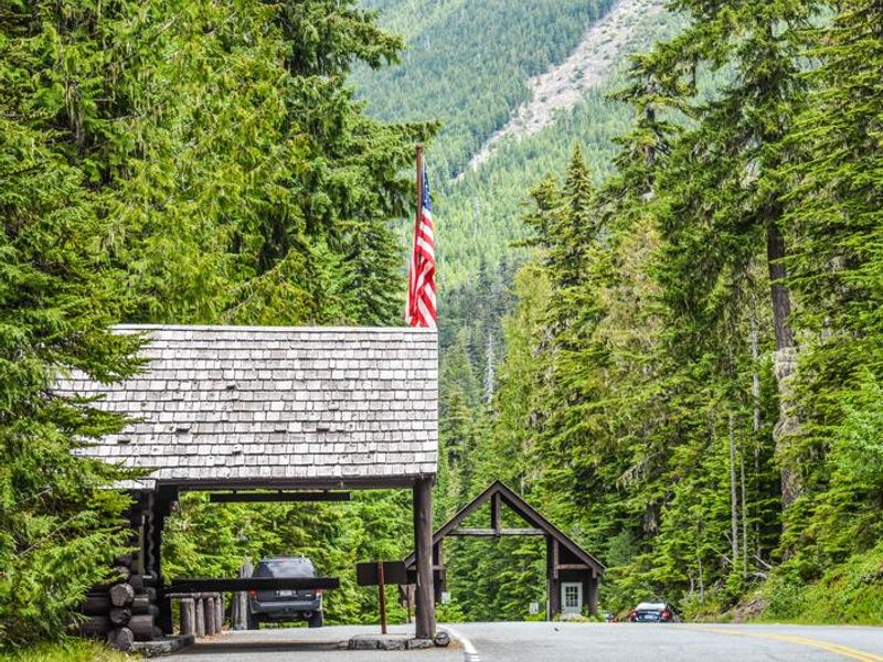 A tree-lined highway with two wooden buildings on the side of the road, one flying an American flag at the White River entrance. Tall conifer trees line the road. A mountain looms in the distance.