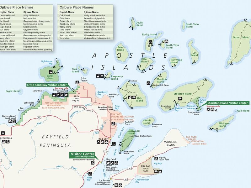 A color image of Apostle Islands National Lakeshore. The islands within the lakeshore are colored in green. The islands have a name on or near them to represent the island name. Some of the islands have dotted lines on them to represent trails. There are two legends on the top left of the image that show the English & Ojibwe names of the islands. 