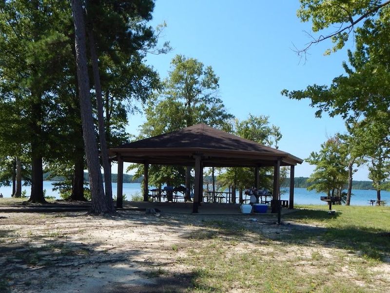This is a picture of the Palmer Point shelter. There are picnic tables under a covered shelter. There is a gravel walkway to the shelter. It has a grill located near the shelter and there are trash cans throughout the park. 