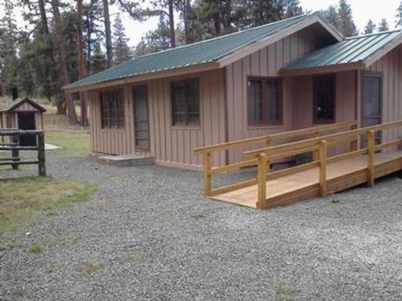 Cabin with beige board-and-batten siding and an entrance ramp in front of an outhouse.