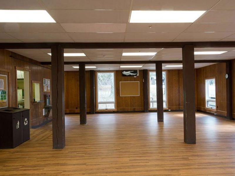 Interior view of dining hall.  Oak colored wood laminate flooring.  Dark brown wooden beams, several large windows, waste/recycle bins and bulletin boards are visible.