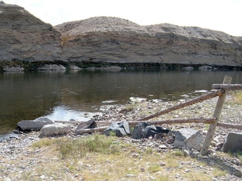 View of a large rock face and a river with a fence made of thin tree trunks on the right.