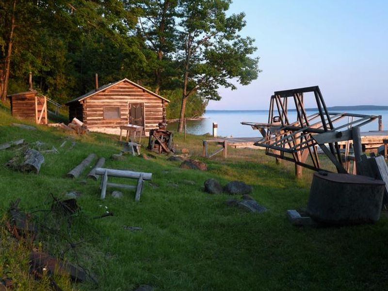 A color photo of the Manitou Fish camping view from the land overlooking the grounds and lake . There is a small building on the far left with a large cabin to the right of it. Trees are behind the buildings. A fishing net reel is on the far right side of the photo with the lake in the background. 