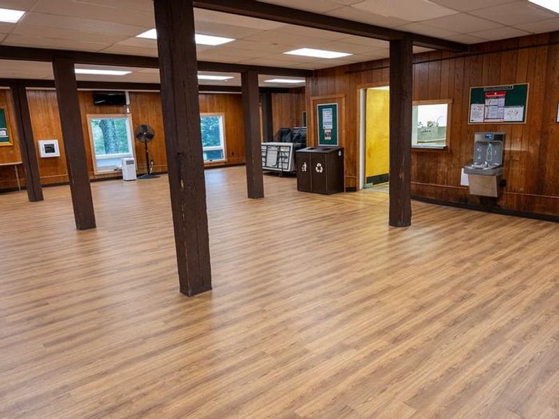 Interior view of dining hall shows 5 large wooden beams, oak laminate flooring, portable a/c unit, pedestal fan, stacked chairs, 6 ft. tables, bottle filling station, waste/recycle bin, and doorway to dishwashing room.