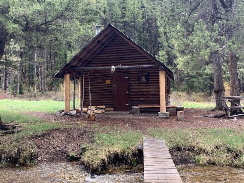 A cabin sets near a creek in a beautiful backcountry setting with a foot bridge over the creek.