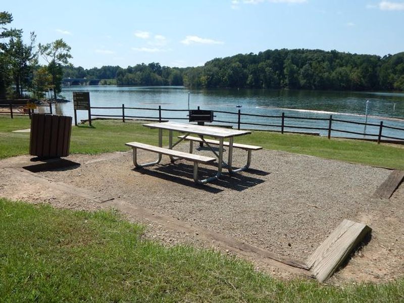 This is a picture of a picnic site at Rudds Creek Day Use Area. This site has timbers around it. There is a picnic table in the graveled area along with a small grill. There is a trash can located to the side of the picnic site. 