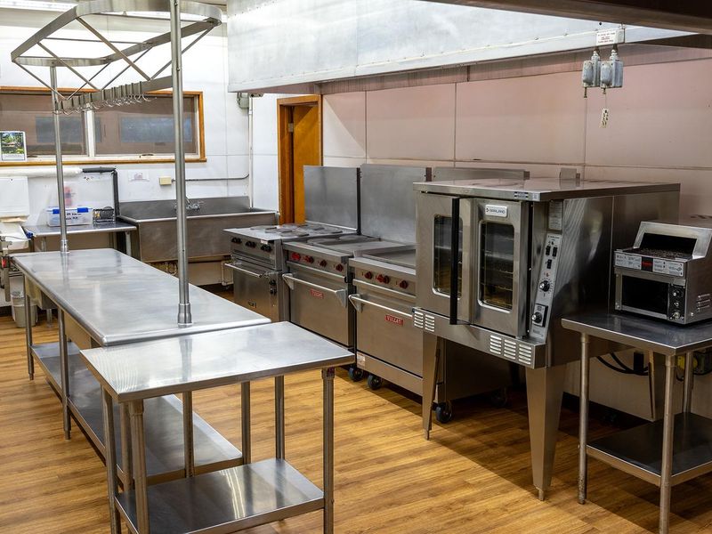 Interior view of the kitchen which is connected to the dining hall. The area has heat and air conditioning. Visible equipment includes stainless prep tables, convection oven, 3 gas ranges, toaster, vent hood and double sink.  